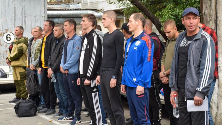 Мужчины разного возраста стоят в очереди у ворот с цифрой 6